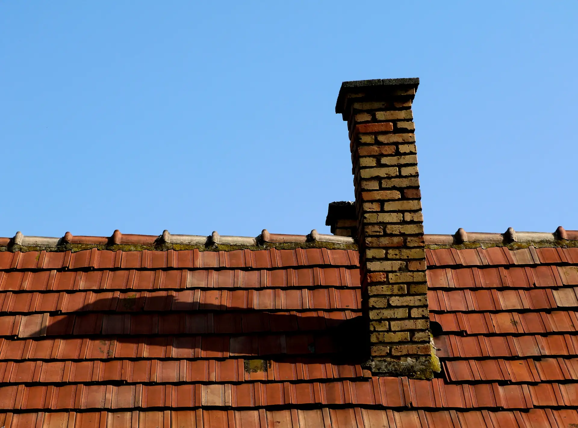 A leaning brick chimney on a tile rooftop against a clear blue sky